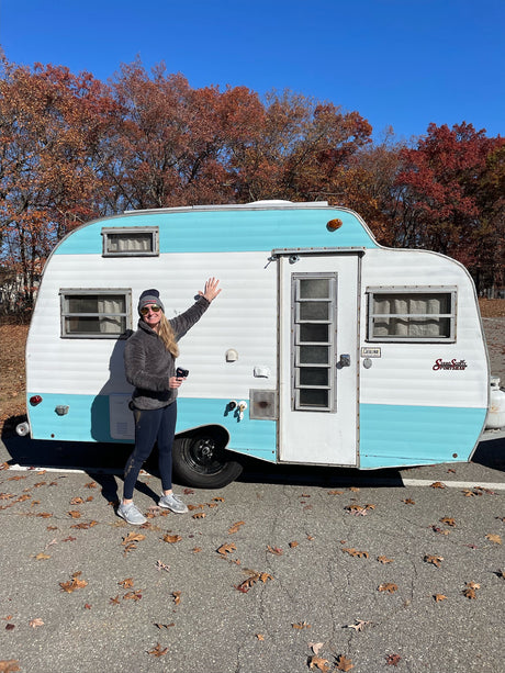 Happy woman waves by a vintage light blue & white Serro Scott Sportsman camper, ideal for an rv rental in autumn.