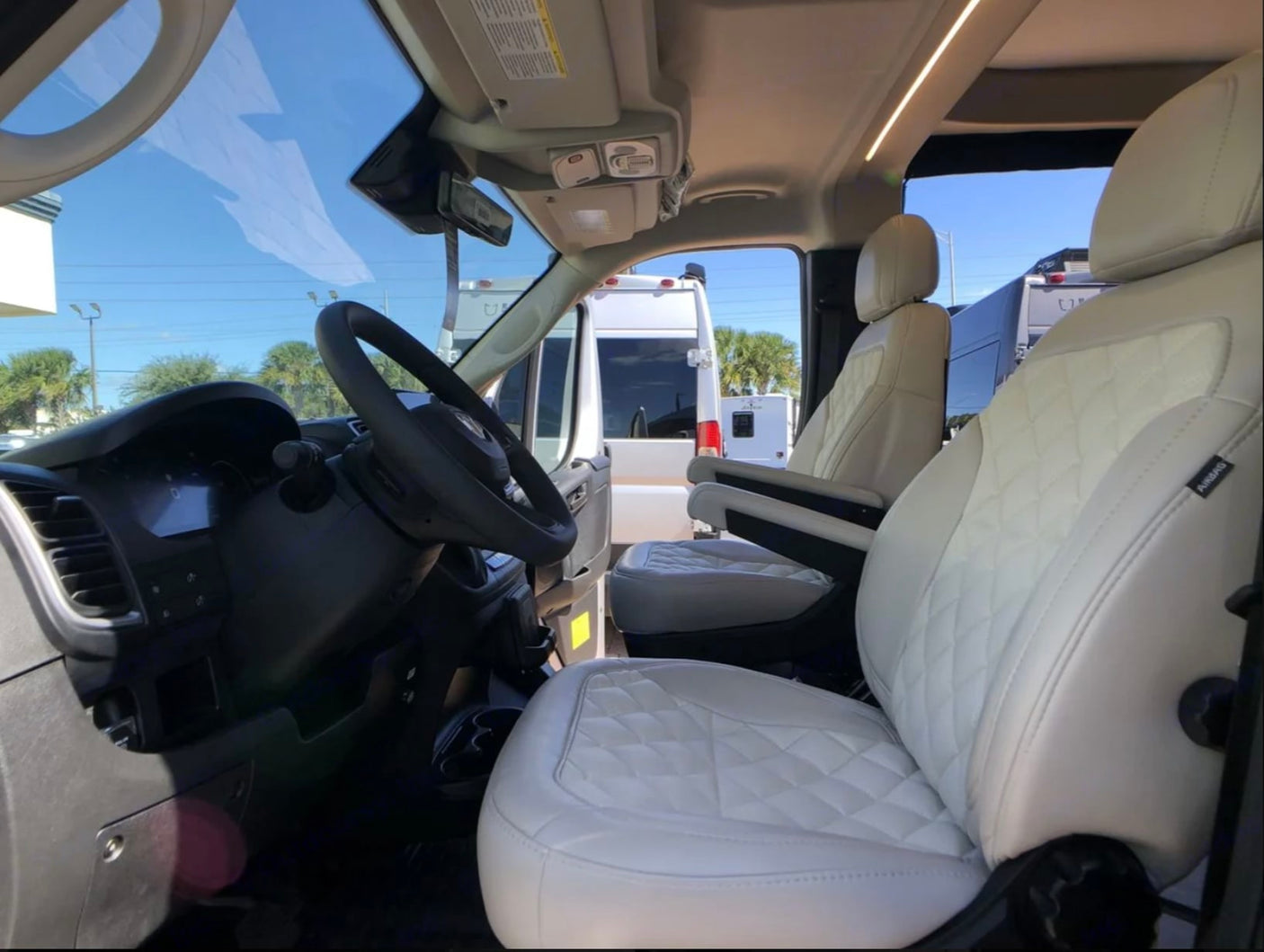Step into this bright RV rental with quilted white front seats, dashboard, and steering wheel, offering views of other RVs outside.