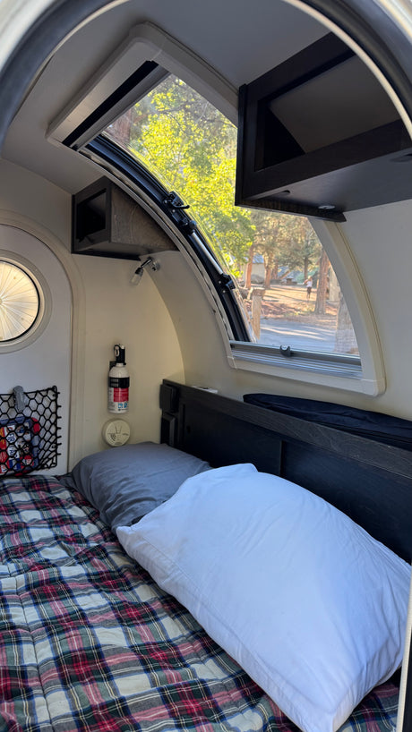 Inside a cozy RV rental: plaid bed, window with forest view, overhead storage, and safety equipment.