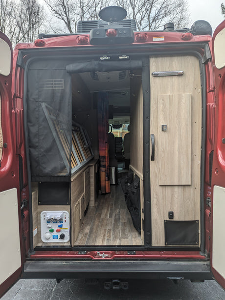 Camper van RV rental: Rear interior view of a red van with open doors, showing kitchen, fold-up bed, & storage.