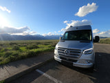 Silver Mercedes Sprinter RV rental parked in a stunning landscape with snowy mountains & green fields.