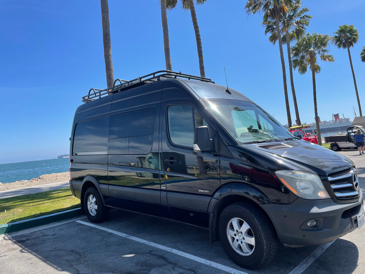Black Mercedes Sprinter RV rental van near Long Beach oceanfront. Sunny day with palm trees, a large ship, and blue sky.