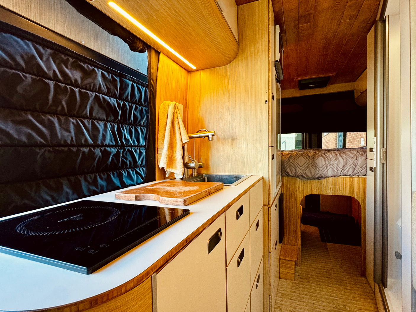 Explore this efficient RV rental with a modern kitchen, induction stove, sink, and comfy raised bed in warm wood tones.