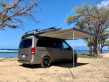 Gray camper van RV rental parked on a sandy beach with its awning extended towards the ocean. Ideal for beach camping & travel.
