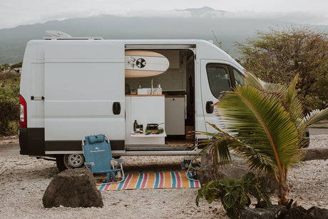 Ideal RV rental for van life adventures! A white camper van with open door reveals a kitchen, surfboard, & beach setup.