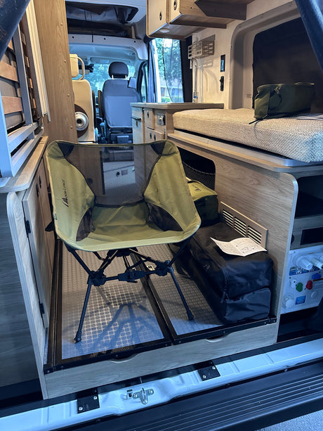 This rv rental interior features a compact, foldable chair on a pull-out tray, next to a bed & kitchenette area.