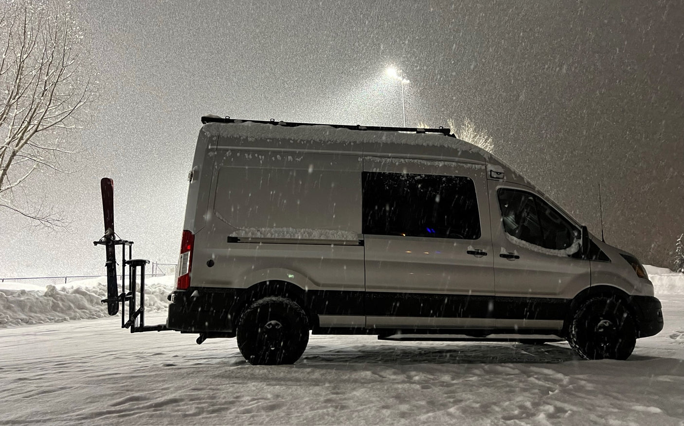 Snow-covered rv rental: White camper van with skis on back in heavy snowfall. Ideal for winter adventures.
