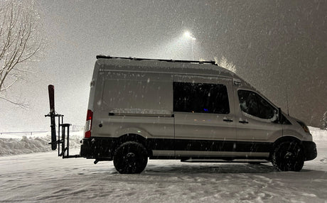 Snow-covered rv rental: White camper van with skis on back in heavy snowfall. Ideal for winter adventures.