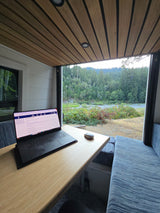 Enjoy remote work in your RV rental! Laptop & mouse on a table overlook a serene river, lush forest. Cozy interior.