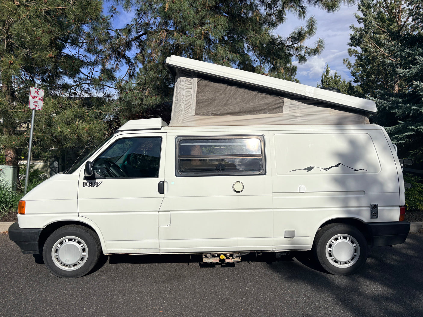 White VW pop-top camper van with its roof extended, parked next to pine trees. This RV rental is ready for journeys.