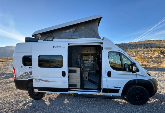 White pop-top Winnebago Solis rv rental, door open to interior. Parked in sunny desert with mountains & clear blue sky.