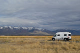 This white rv rental rests amidst golden fields, framed by majestic snow-capped mountains under a dramatic sky.