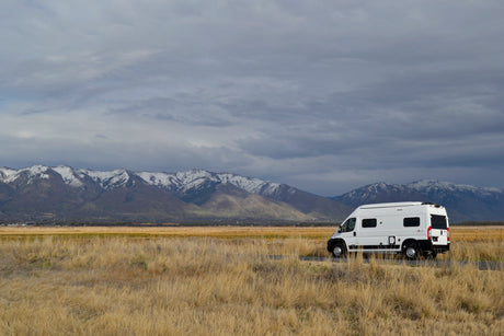 This white rv rental rests amidst golden fields, framed by majestic snow-capped mountains under a dramatic sky.