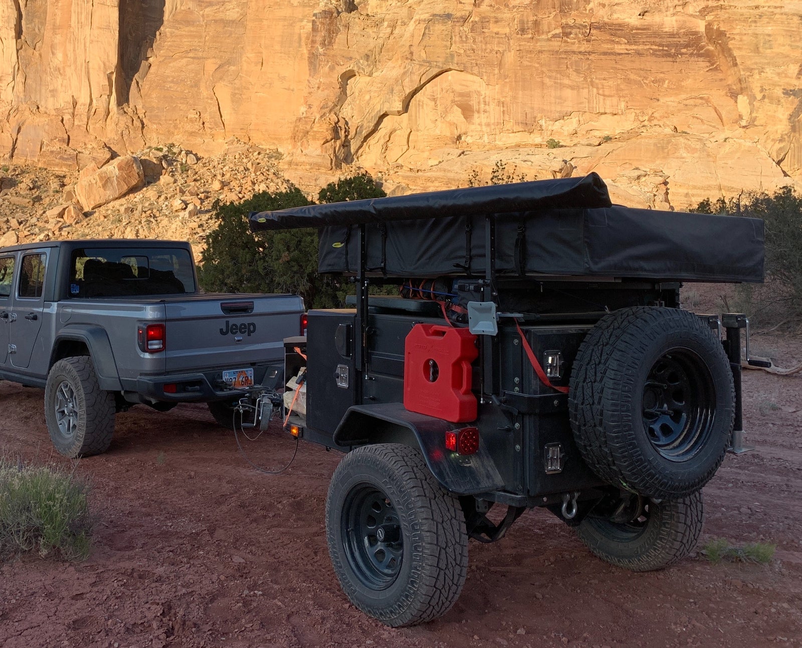 Gray Jeep Gladiator towing an off-road overland trailer with rooftop tent by desert cliffs. Perfect for adventure **RV rental**.