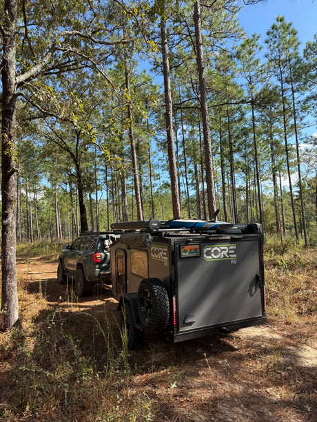 Off-road rv rental adventure: SUV towing a CORE RV trailer with a dog through a serene pine forest.