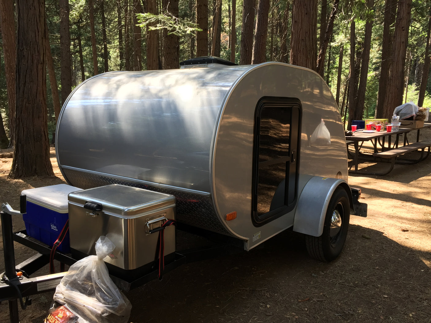 Silver teardrop RV rental at a sunny forest campsite, with a picnic table and gear nearby.