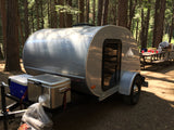 Silver teardrop RV rental at a sunny forest campsite, with a picnic table and gear nearby.