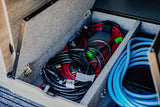 RV rental storage: an open compartment holds coiled red and black electrical cables, and a bright blue water hose.