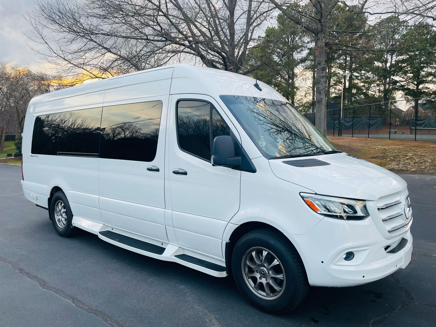  is parked on a gravel lot with trees and old structures behind it.|White Mercedes Sprinter van. A spacious rv rental parked among sunlit trees — photo 3