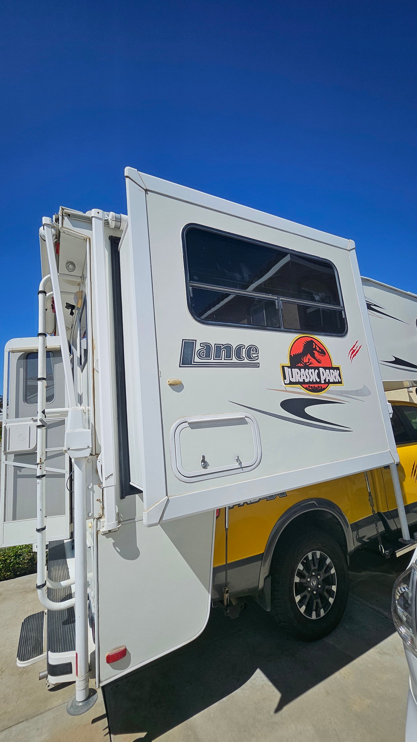 A white Jurassic Park themed Lance truck camper, ready for adventure as an RV rental, parked under a clear blue sky.