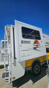 A white Jurassic Park themed Lance truck camper, ready for adventure as an RV rental, parked under a clear blue sky.