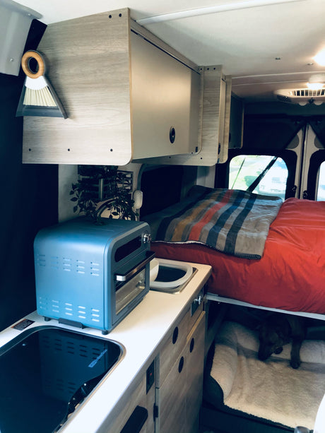 This cozy RV rental interior features a compact kitchenette, bed with red linens, overhead storage, and a dog's bed below.