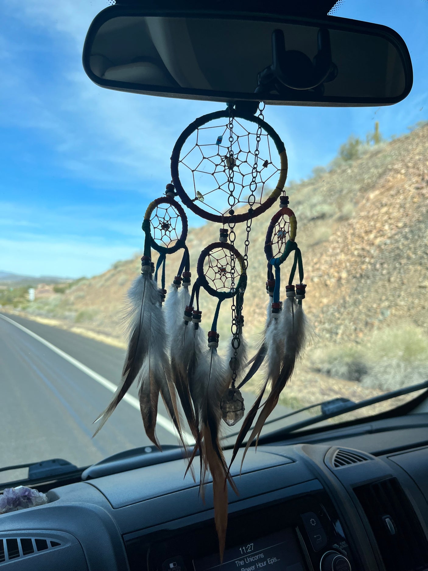 A dreamcatcher with feathers and crystals hangs from the rearview mirror of an RV rental, on a scenic desert road trip.