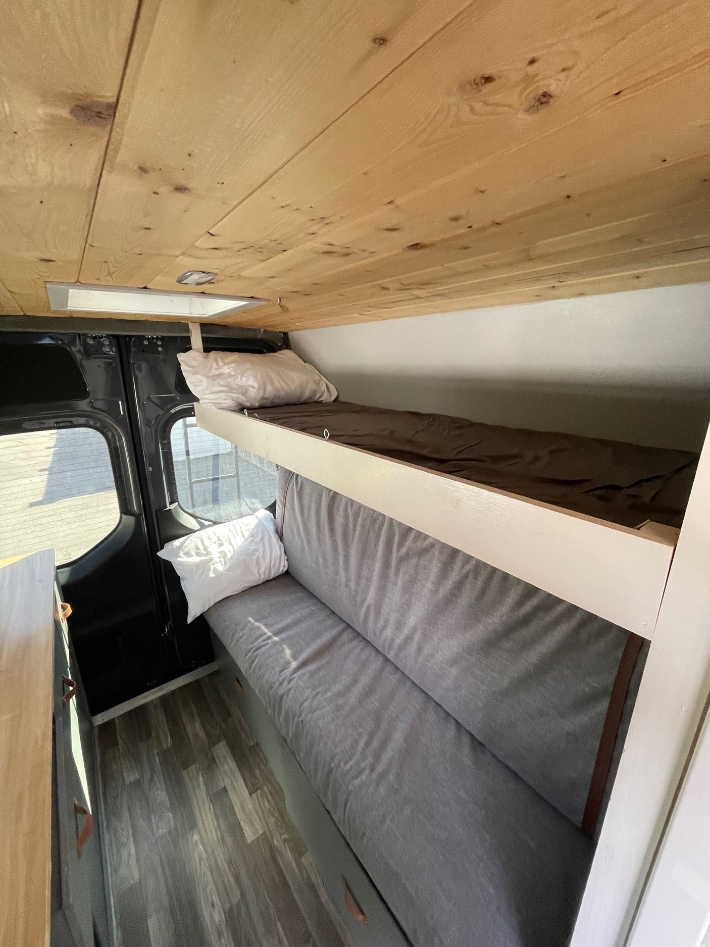 Cozy rv rental interior with wooden ceiling, featuring bunk beds (upper with brown sheet, lower grey bench) and laminate floor.