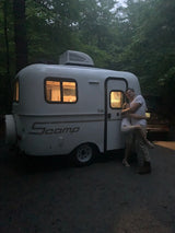 A white Scamp compact RV rental glows at dusk, with a person standing on its side. Ideal for outdoor adventures.