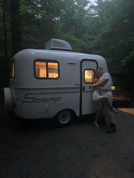 A white Scamp compact RV rental glows at dusk, with a person standing on its side. Ideal for outdoor adventures.
