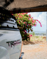 Close-up of an adventure-ready TRD Off Road truck, likely an rv rental, framed by a dirty roof, overlooking a bay with pink flowers.