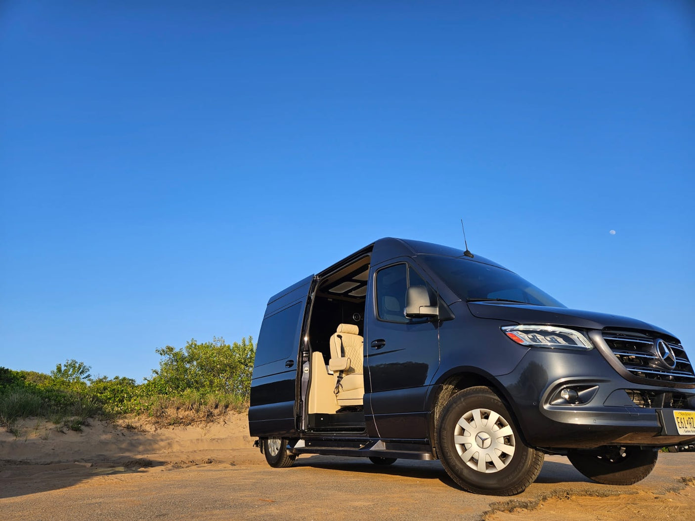Experience premium travel with our luxury Mercedes-Benz Sprinter RV rental, featuring an open side door revealing a plush interior on a sandy road.