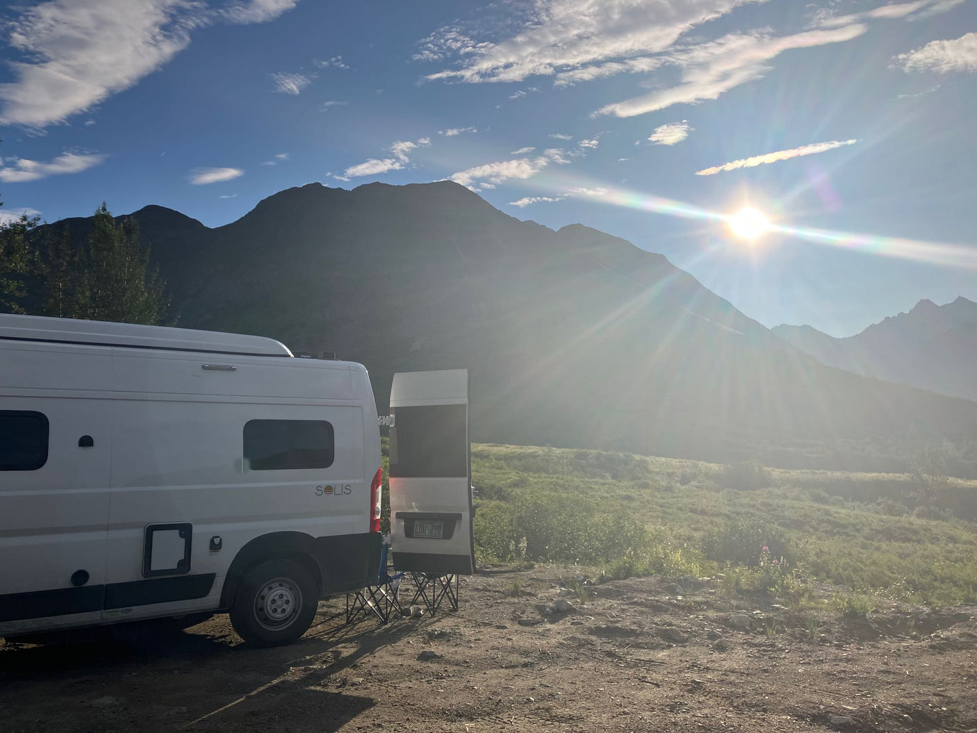 Scenic RV rental adventure. A white camper van is parked amidst sunny mountains and a green field under a bright blue sky.