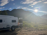 Scenic RV rental adventure. A white camper van is parked amidst sunny mountains and a green field under a bright blue sky.