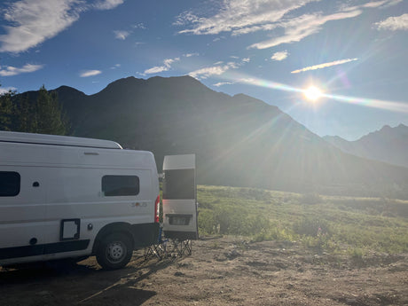 Scenic RV rental adventure. A white camper van is parked amidst sunny mountains and a green field under a bright blue sky.