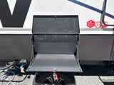 Open black metal storage compartment on an RV, revealing inner shelves, utility hookups, and a tire. Useful for RV rental setup.