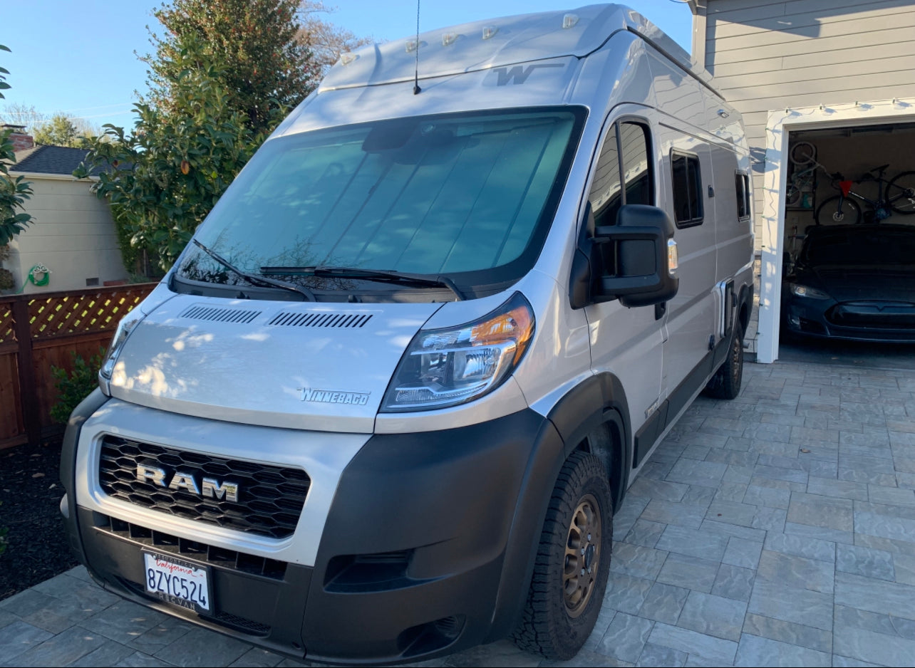 Silver Winnebago Ram ProMaster camper van for rv rental, parked on a driveway with a Tesla in the garage.