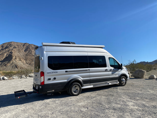 Silver rv rental van with cargo carrier parked on gravel, surrounded by desert mountains under a clear blue sky.