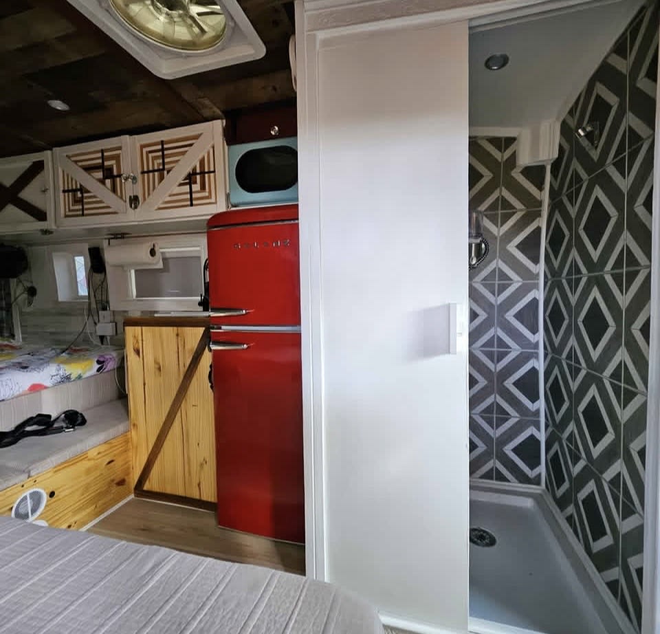 This RV rental's compact interior includes a red retro fridge, microwave, wood cabinets, a shower with stylish patterned tiles, and a cozy bed/seating.