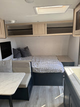 Modern RV rental interior with a cozy bed, dinette, overhead storage, and kitchen counter. Bright and functional for travel.