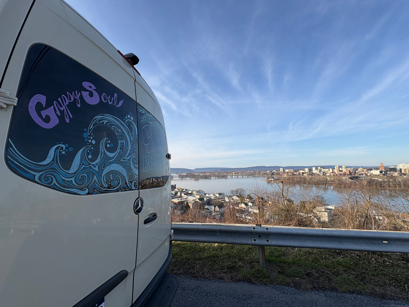 This 'Gypsy Soul' rv rental camper van, adorned with wave art, offers a stunning view of a city & river against a blue sky.