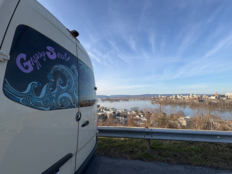 This 'Gypsy Soul' rv rental camper van, adorned with wave art, offers a stunning view of a city & river against a blue sky.