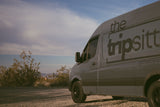 A gray "the tripsitter" RV rental van is parked on a dusty desert road, surrounded by sparse bushes under a partly cloudy sky.