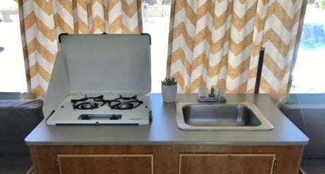 Compact RV rental kitchenette: a gas stove, sink, potted plant, and chevron curtains. Perfect for cooking on the go.