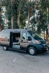 Gray camper van RV rental with side door open, revealing comfortable interior. Parked by trees, ready for your road trip.
