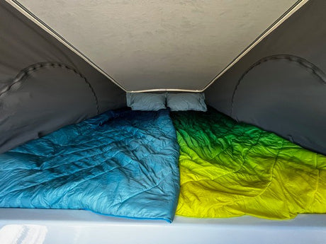 Inside a pop-top RV rental: a cozy bed with bright blue & green-yellow comforters and pillows. Ready for adventure.