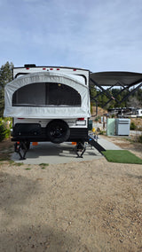 Back of an RV rental pop-up travel trailer with extended bed, awning, and leveling jacks at a campsite. Perfect for camping.