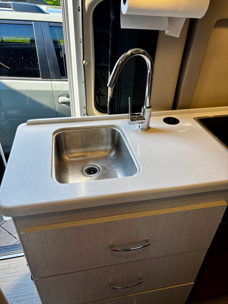 RV rental kitchen: modern galley with stainless steel sink, chrome faucet, speckled countertop, and light wood-grain drawers.
