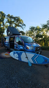 Blue/purple custom RV rental camper van with pop-top tent & paddleboard on gravel. Perfect for outdoor adventures!