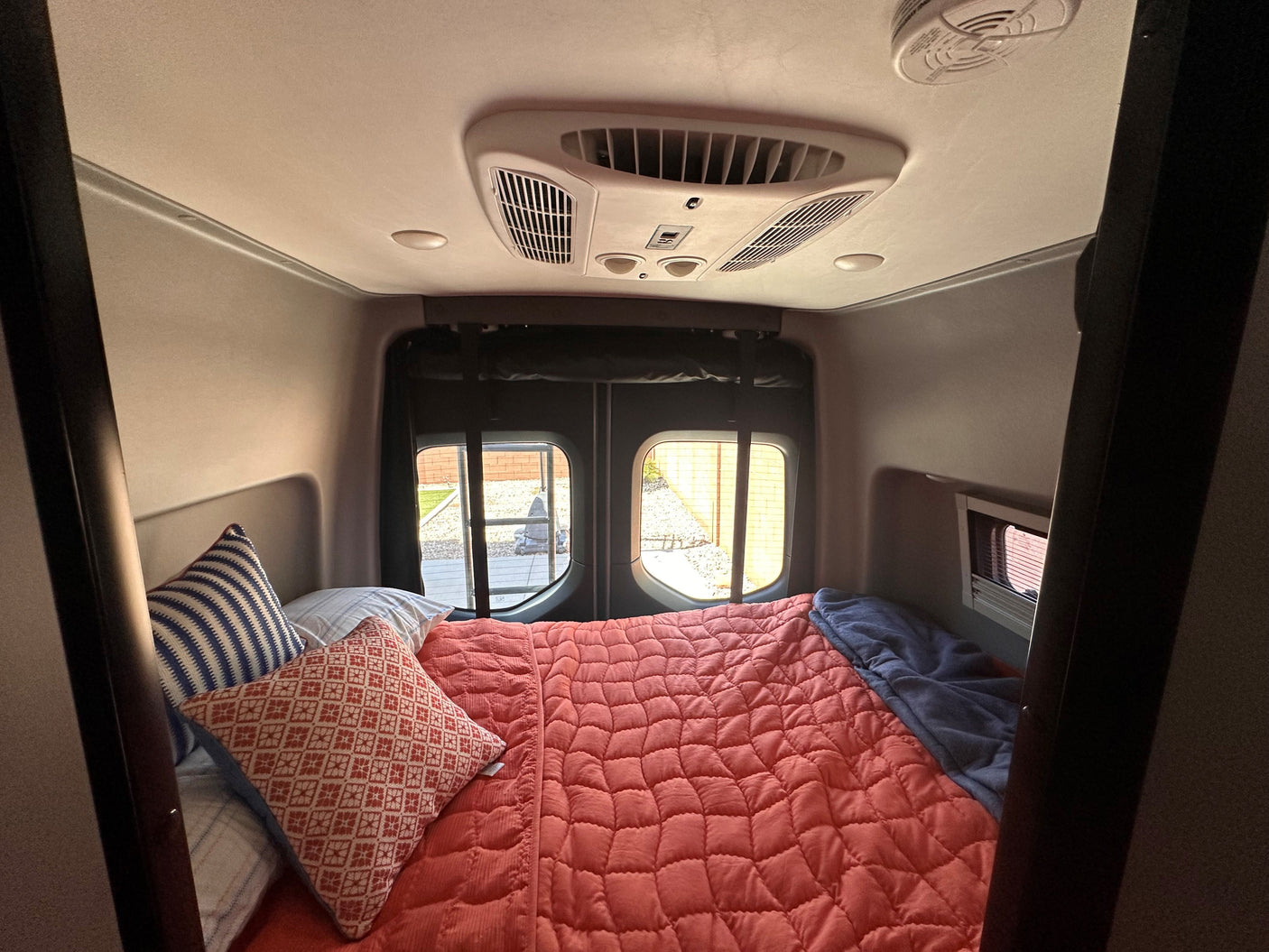 Comfortable RV rental bed setup with a red comforter and pillows. Rear windows show outdoor view; AC unit on ceiling.